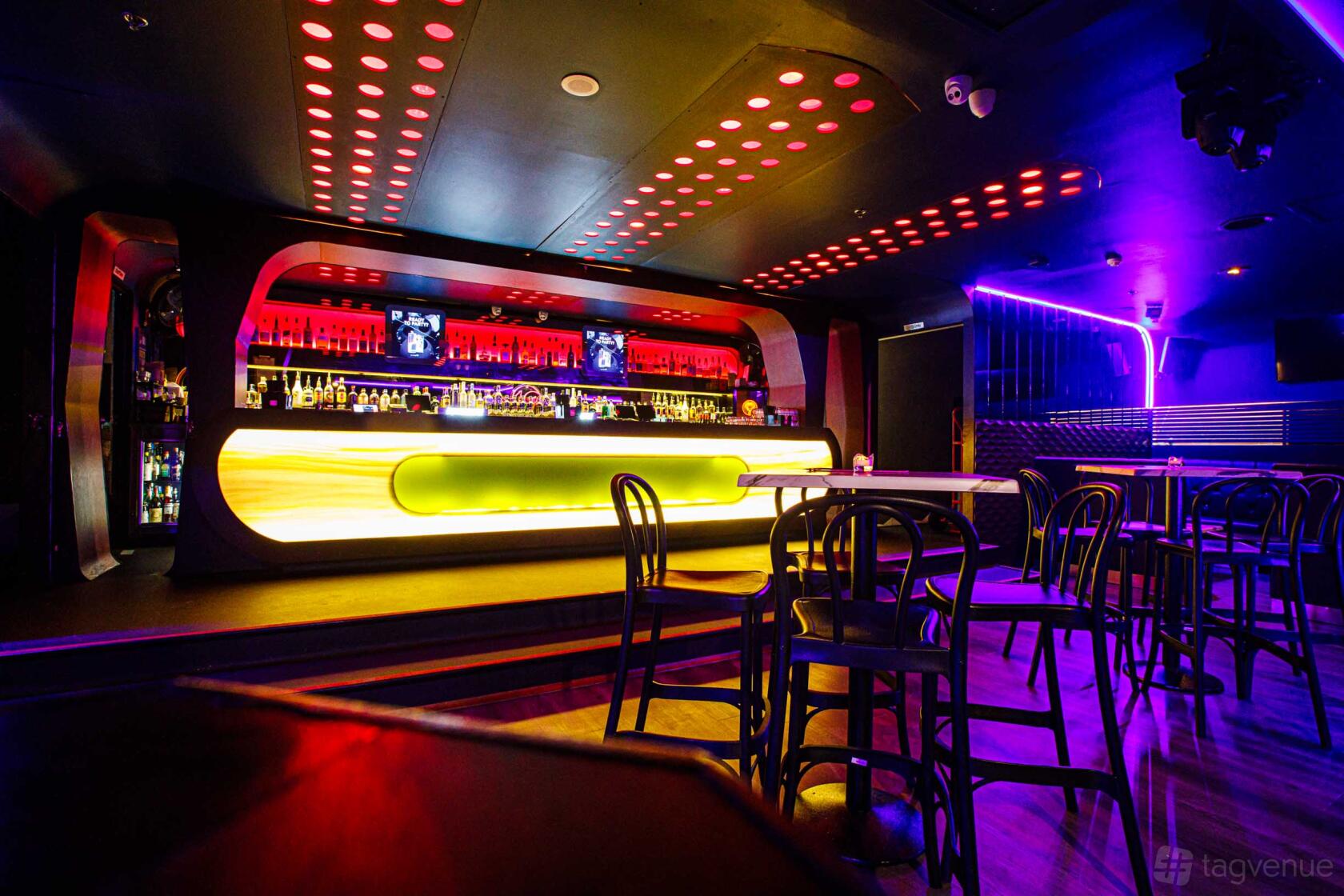 A nightclub with a neon-lit bar, glowing countertop, and scattered tables with black chairs at Khokolat Bar.