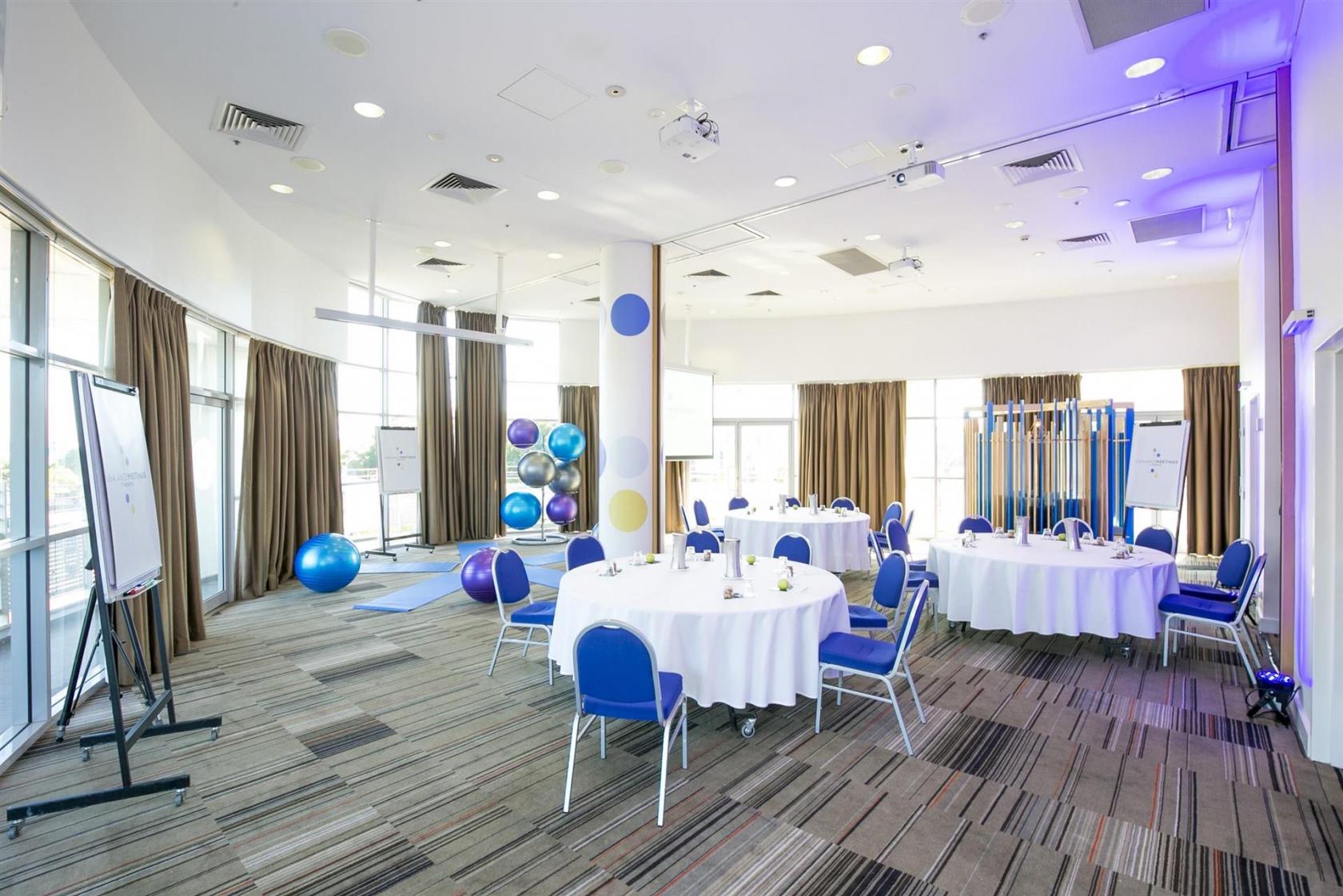 A meeting room with round tables, blue chairs, floor-to-ceiling windows, and exercise balls at Novotel Sydney Olympic Park Hotel.