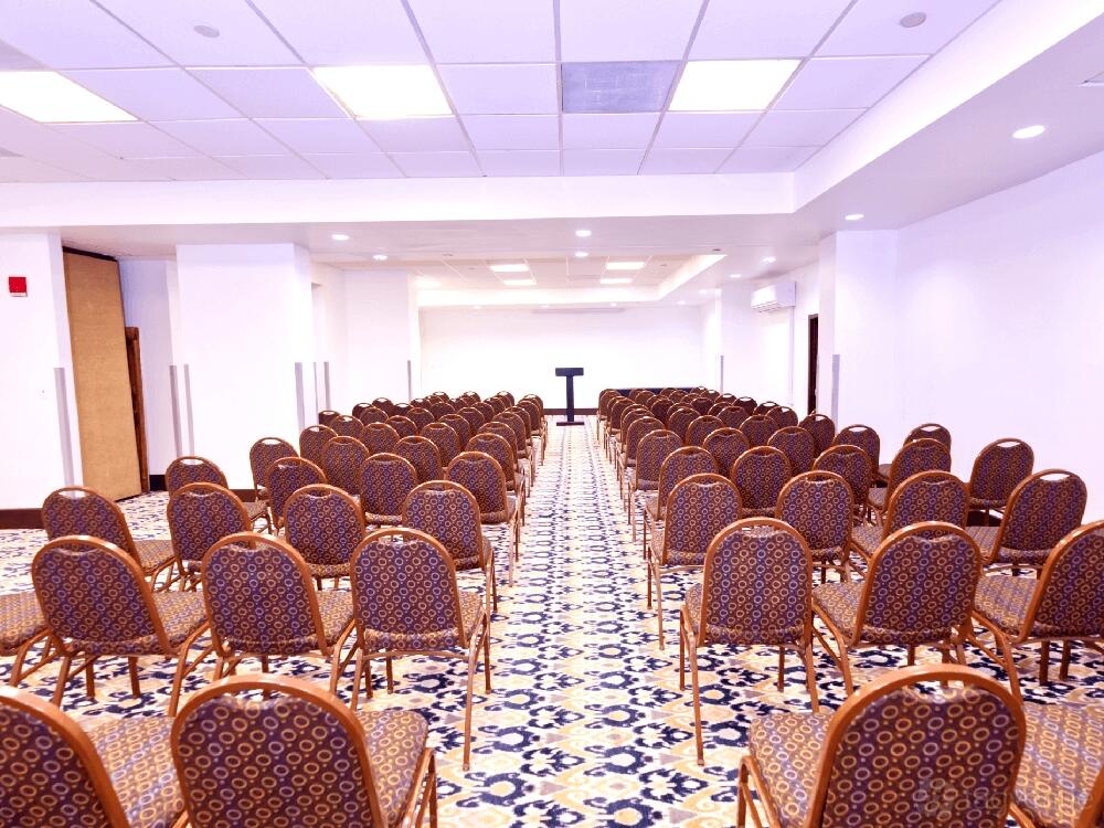A hotel meeting room with patterned carpet, rows of cushioned chairs, and a podium at Harmony Suites Secaucus Meadowlands.