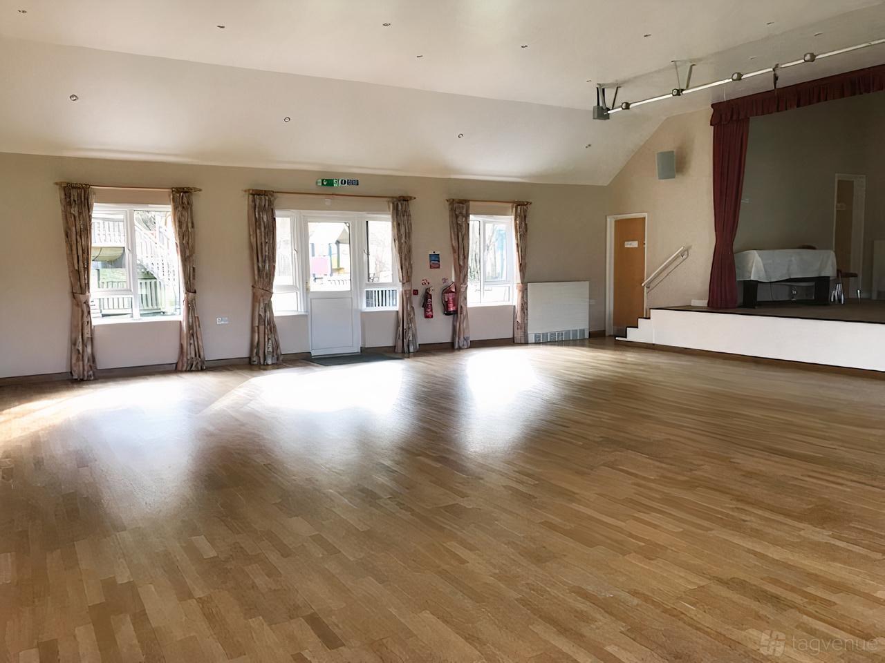 A spacious hall with light wood floors, large windows with curtains, and a small stage at Hascombe Village Hall.
