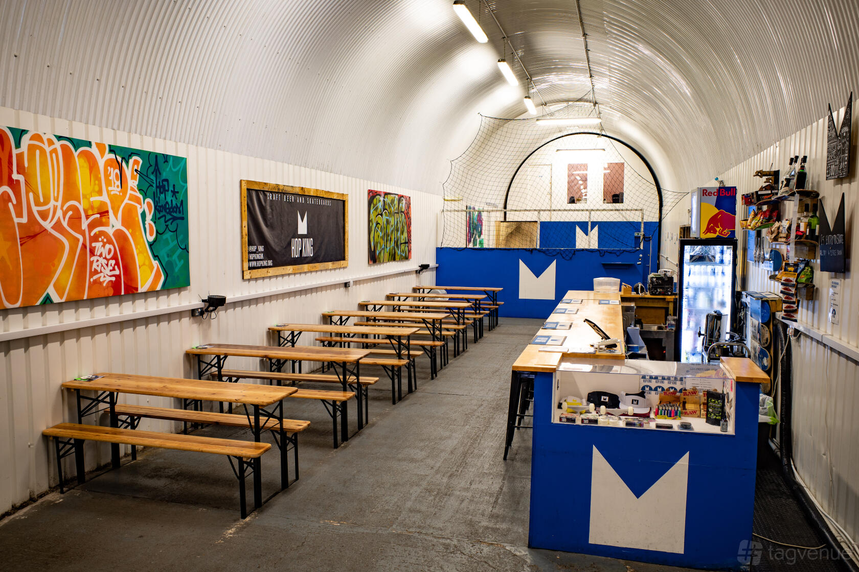 An event venue with long wooden benches, colorful wall art, and an arched metal ceiling at Hop Kingdom.