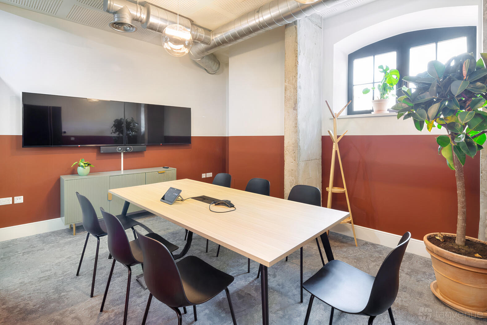 A meeting room with a long wooden table, black chairs, wall-mounted screens, and potted plants at Riley Studios by Spacemade.