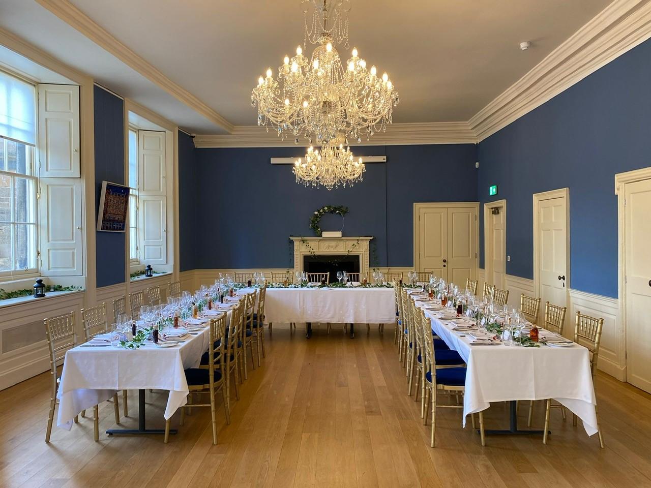 A function room with blue walls, crystal chandeliers, banquet tables with white linens, and large windows at Riddle's Court.