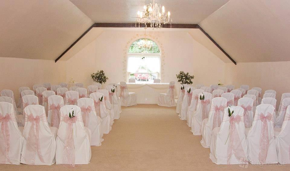 An event space with white chair covers, pink sashes, a chandelier, and a large arched window at Fox & Hounds.