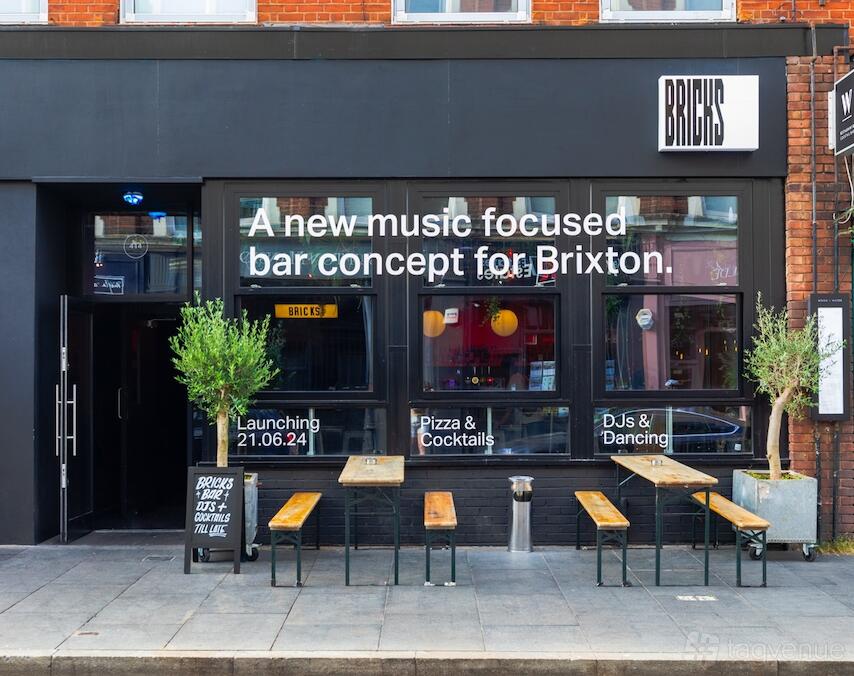 A nightclub exterior with black facade, large windows, wooden benches, and potted trees at Bricks.
