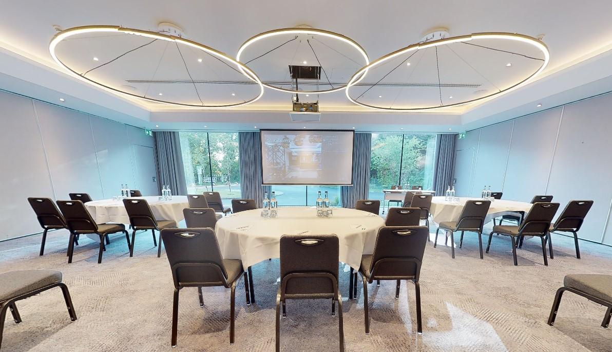 A hotel meeting room with round tables, black chairs, floor-to-ceiling windows, and a projector screen at Holiday Inn Shepperton.