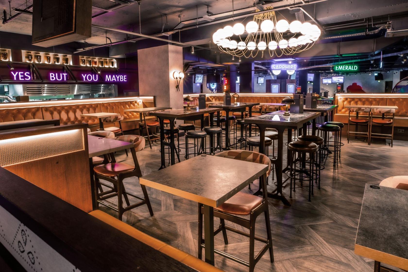 An event venue with high-top tables, leather banquette seating, and a modern chandelier at Puttshack Watford.