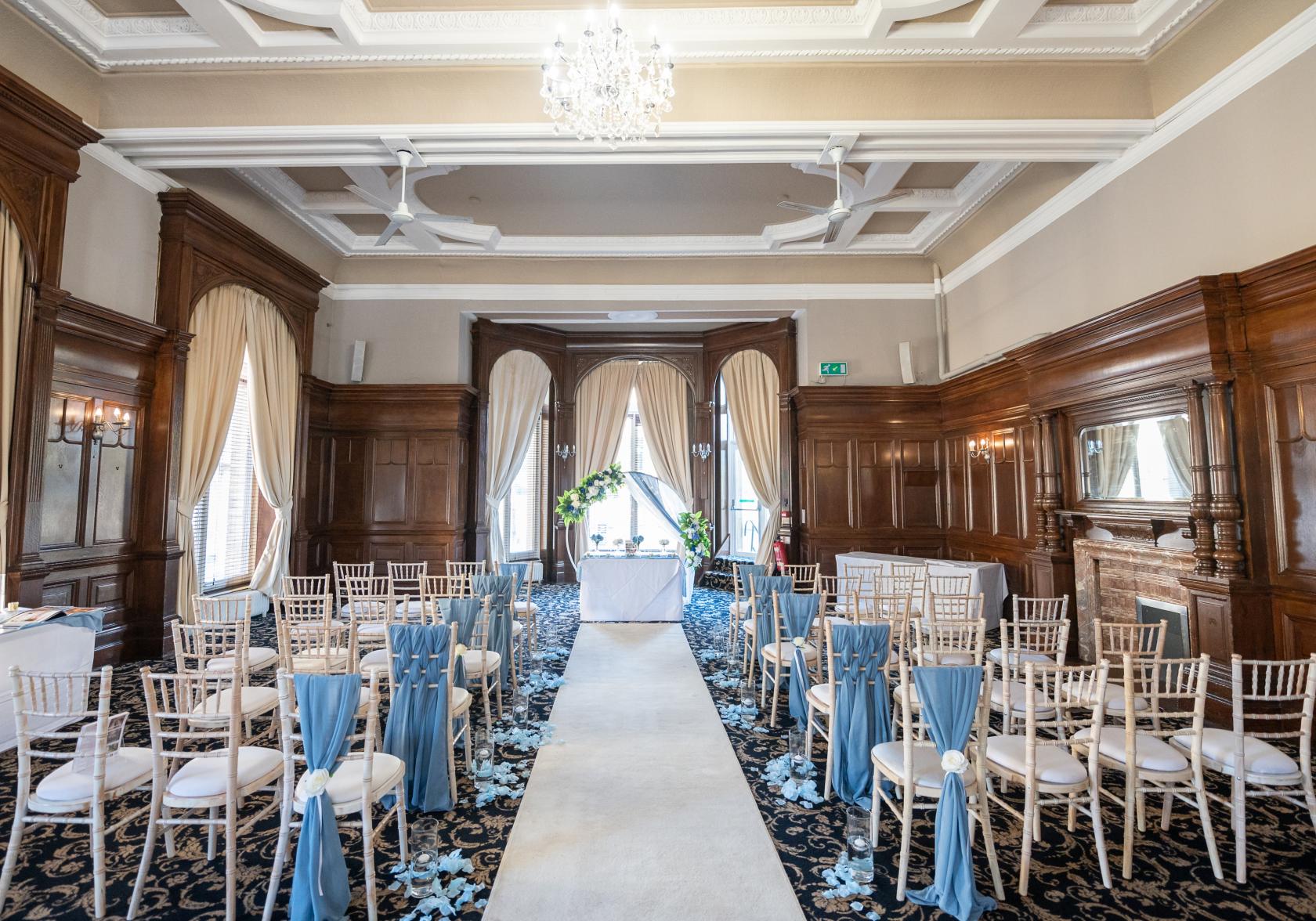 A function room with wood-paneled walls, chandeliers, large arched windows, and an aisle with white chairs at The Great Victoria Hotel