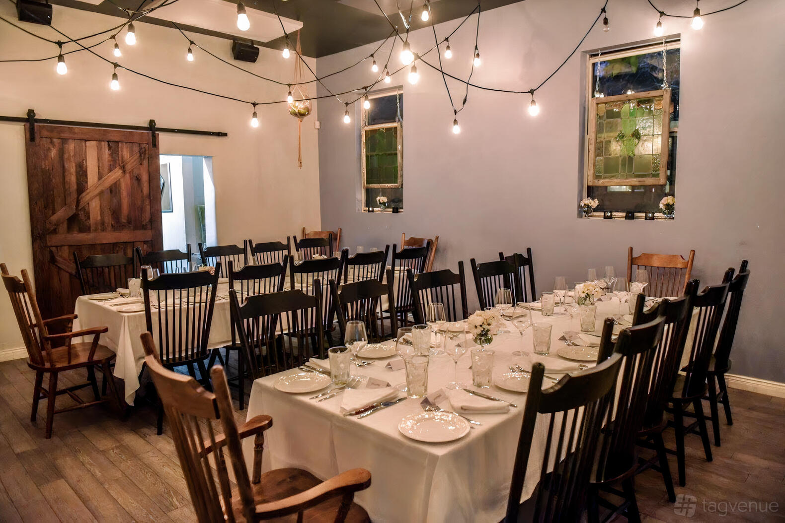 A dining room with wooden chairs, string lights, and two banquet tables set with white linens at Her Father's Cider Bar + Kitchen.