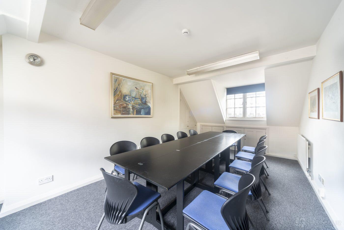 A meeting room with a long black table, blue chairs, framed artwork, and natural lighting at Hellenic Centre.