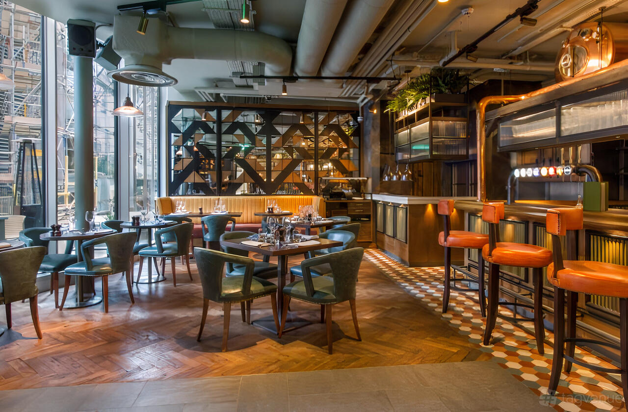 A bar in a pub with geometric wall panels, high ceilings, leather barstools, and wooden flooring at Broadwood.