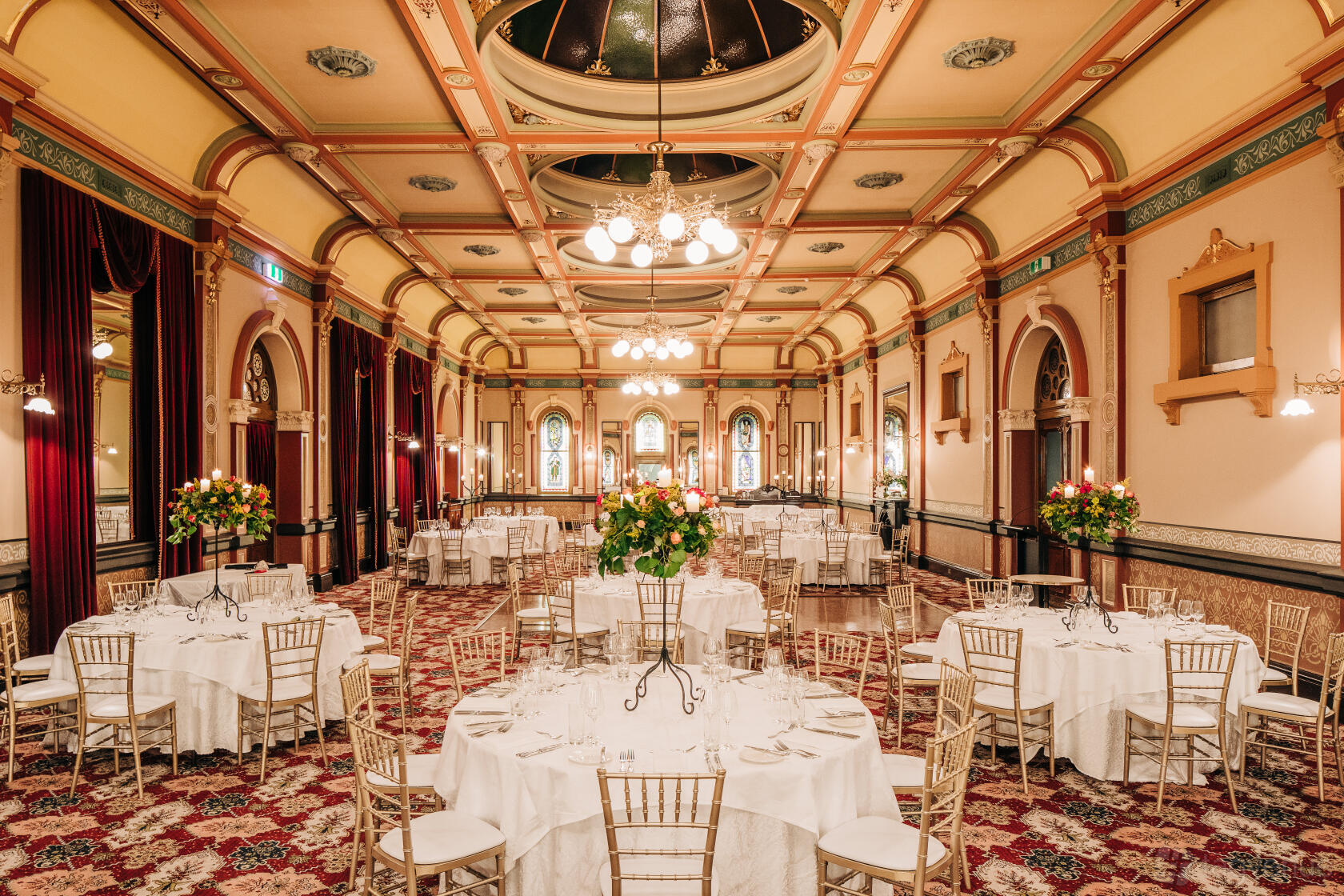 A hotel ballroom with ornate ceilings, chandeliers, arched windows, and round tables with white linens at The Hotel Windsor.