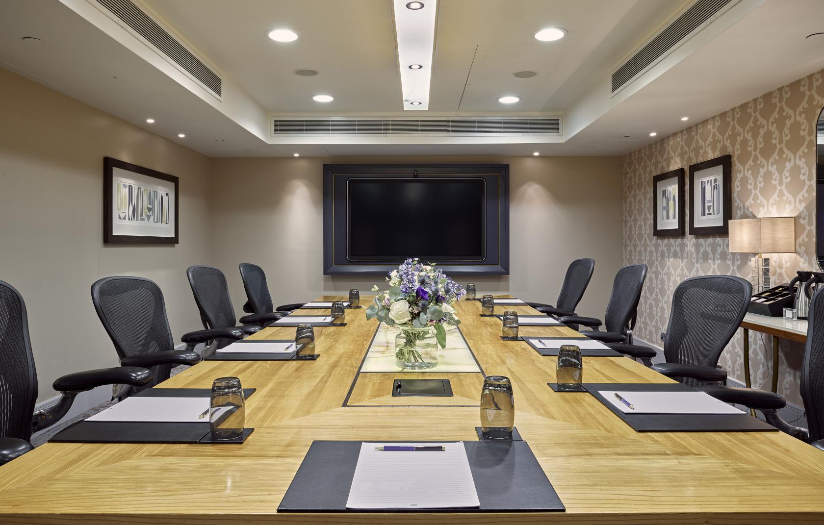 A meeting room with a large wooden conference table, ergonomic chairs, and wall-mounted display at Meetings & Events Venues.