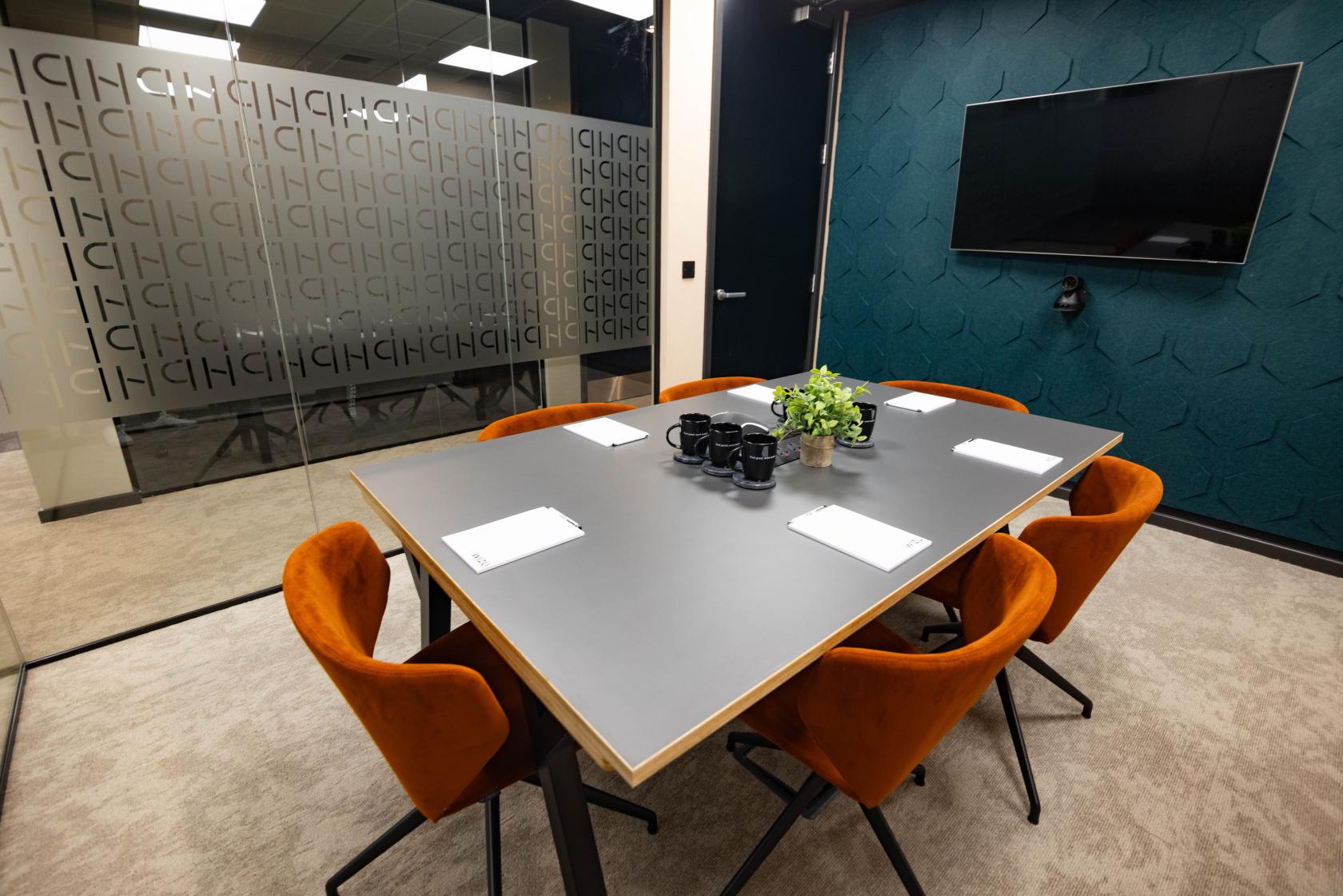 A meeting room with orange chairs, a rectangular table, glass walls, and a wall-mounted screen at Wizu Workspace - Newcastle - Portland House.