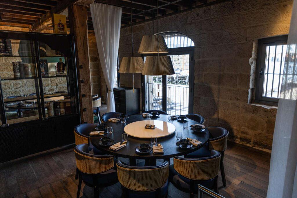 A dining room with a round table set for eight, cane-back chairs, exposed stone walls, and pendant lighting at Bar Lulu.