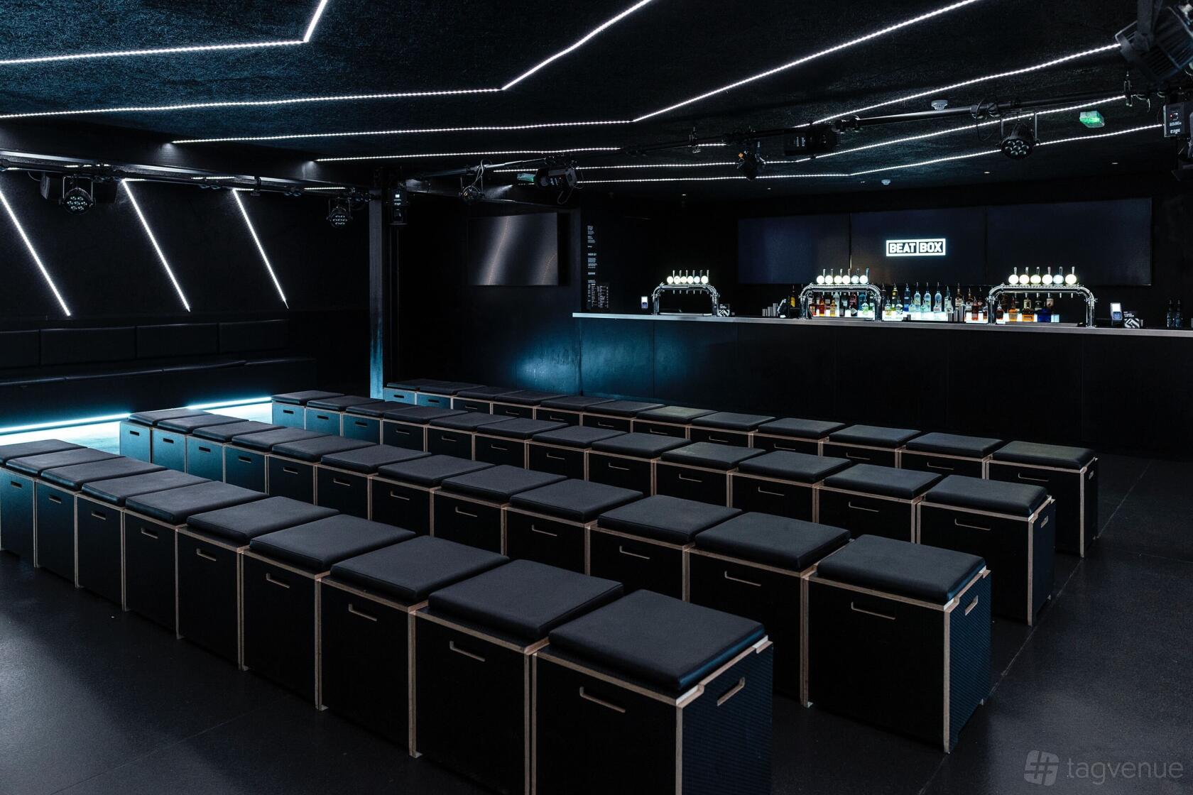 An event space with black cushioned seating, a backlit bar, and geometric ceiling lights at Boxpark Wembley.