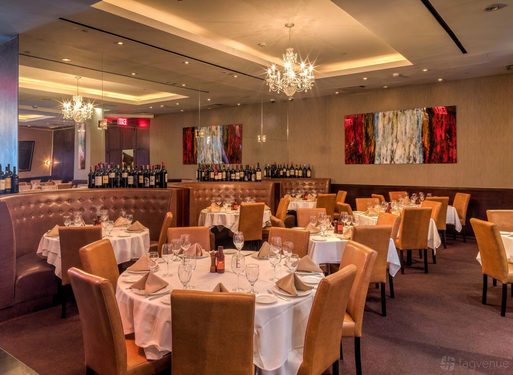 A restaurant dining room with tan upholstered chairs, round tables with white linens, chandeliers, and modern art at Empire Steak House West.