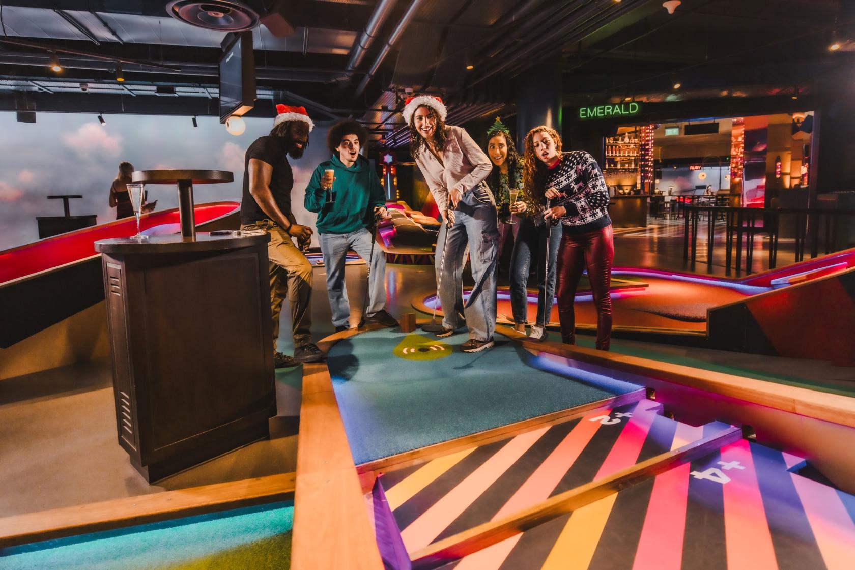 An event venue with neon-lit mini golf courses, colorful flooring, and a modern bar area at Puttshack Bank.