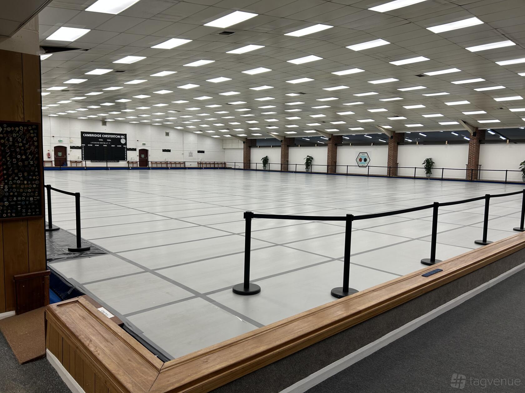 A sports hall with a large indoor bowls arena, bright overhead lighting, and surrounding seating at Cambridge Chesterton Indoor Bowls Club.
