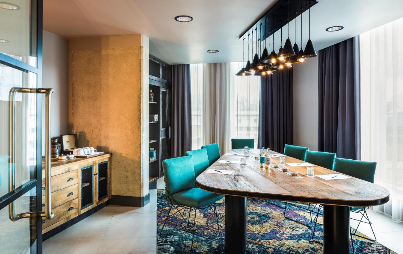 A meeting room with a large wooden table, teal chairs, pendant lighting, and floor-to-ceiling windows at Novotel Canary Wharf.