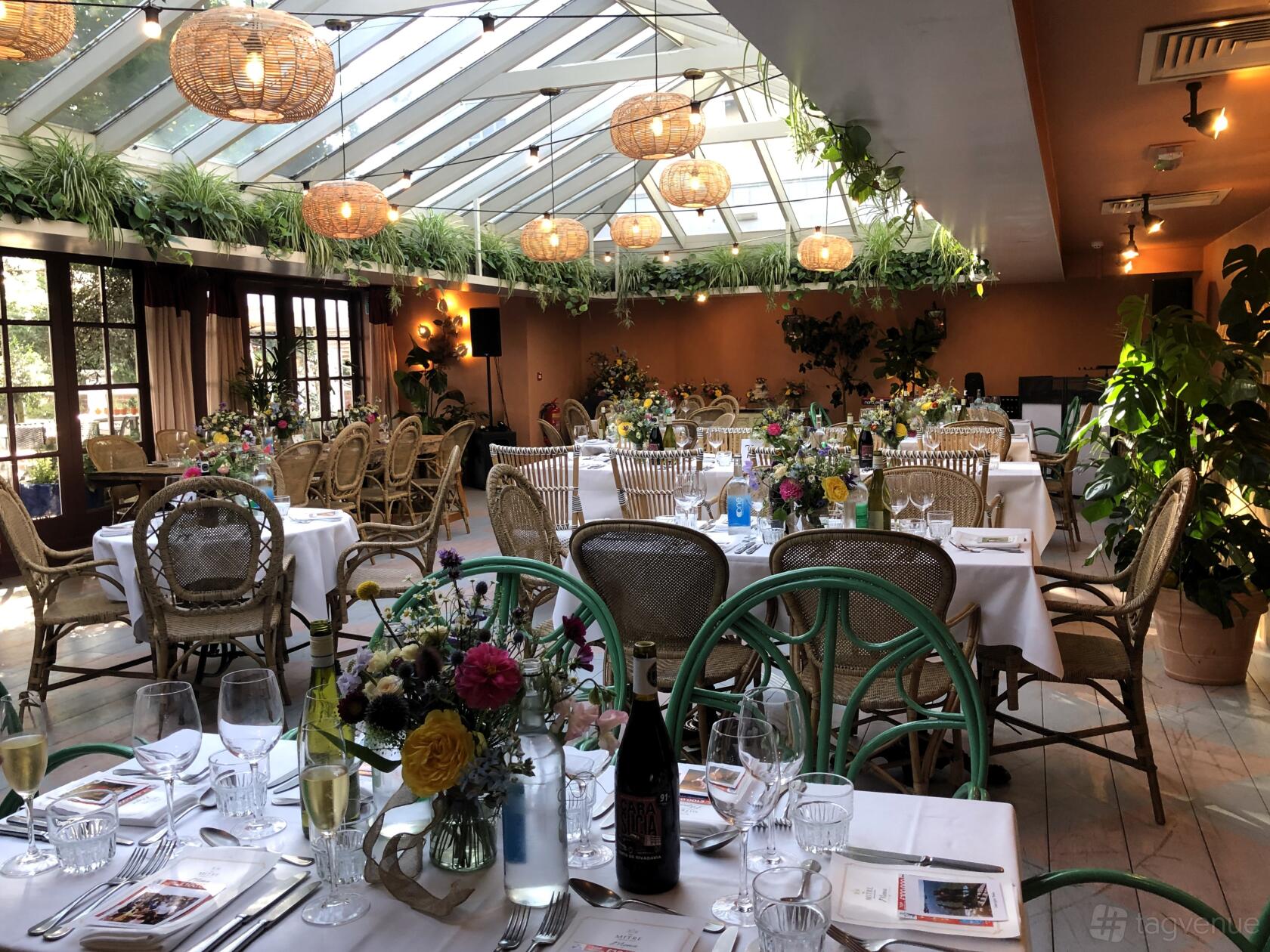 A function room with a glass ceiling, rattan chairs, and floral table arrangements at The Mitre.