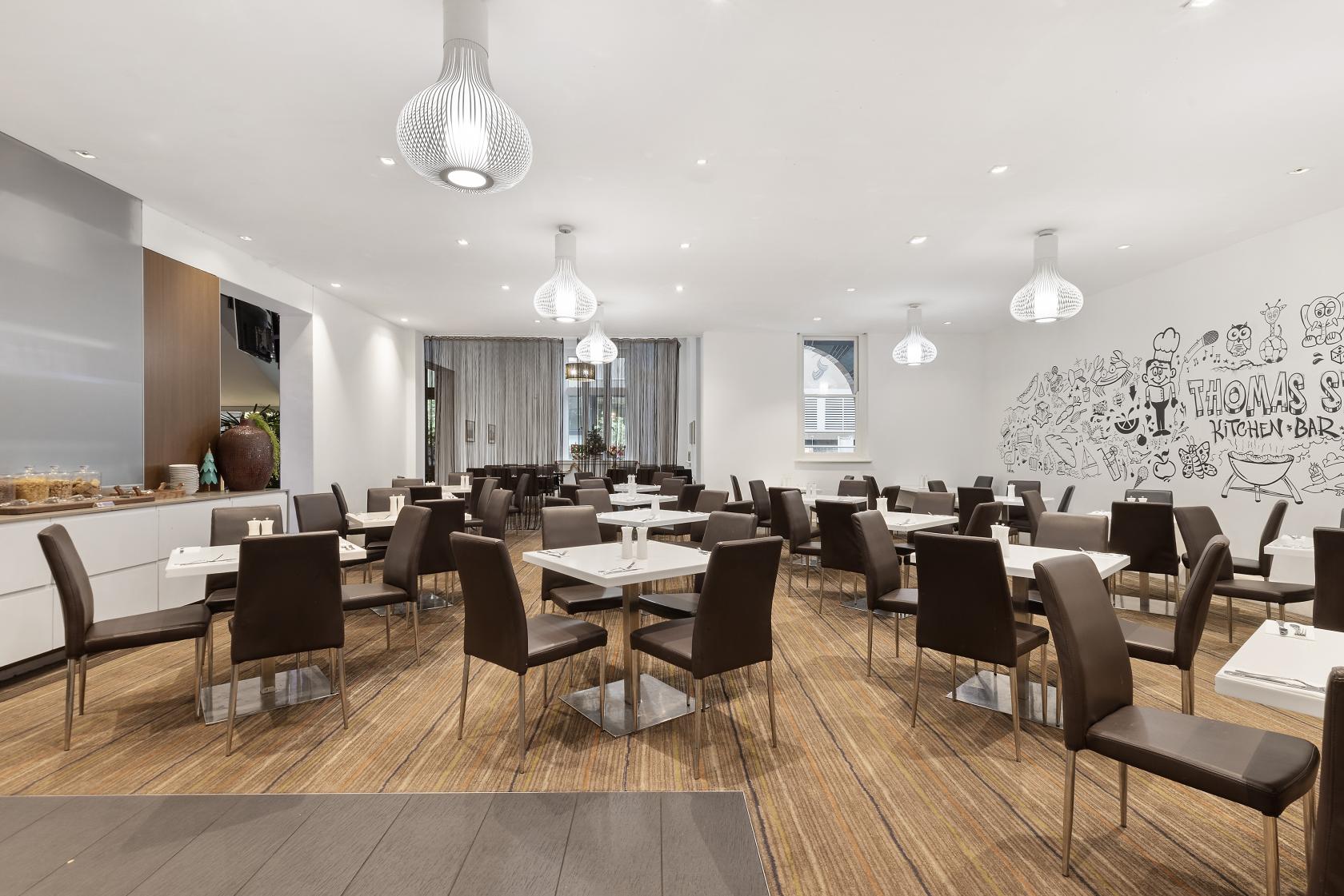 A hotel restaurant with dark brown chairs, white square tables, pendant lighting, and a wall mural at Sydney Central Hotel.