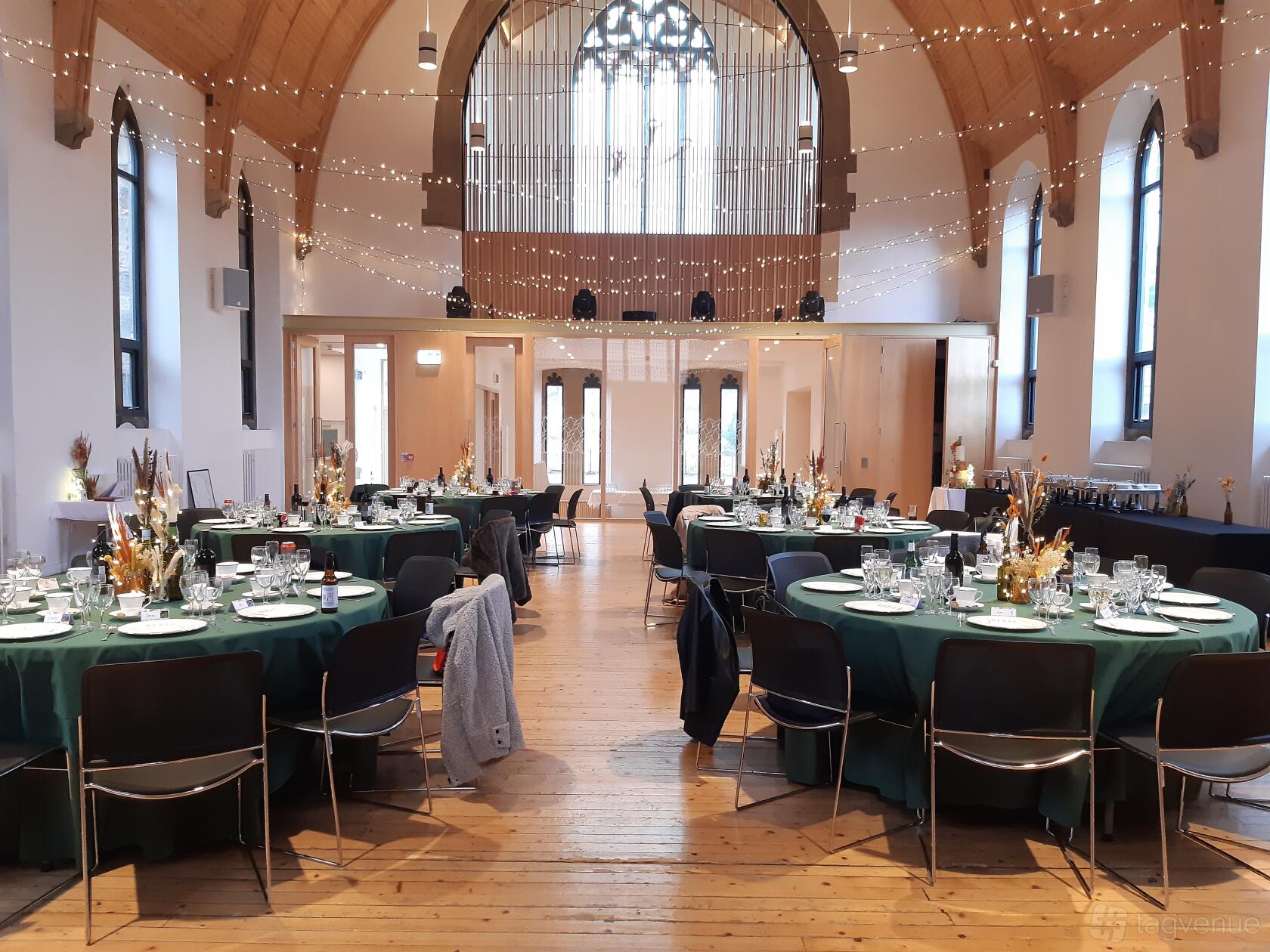 A community hall with round tables set with green linens, glassware, string lights, and arched windows at Greyfriars Charteris Centre.