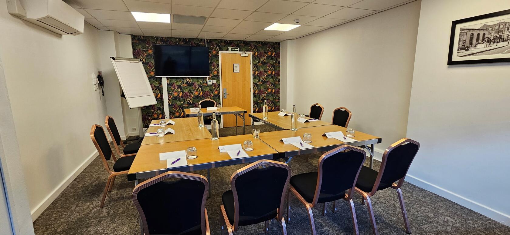A meeting room with a rectangular table setup, black chairs, flip chart, wall-mounted screen, and patterned accent wall at Manchester Conference Centre & Pendulum Hotel.