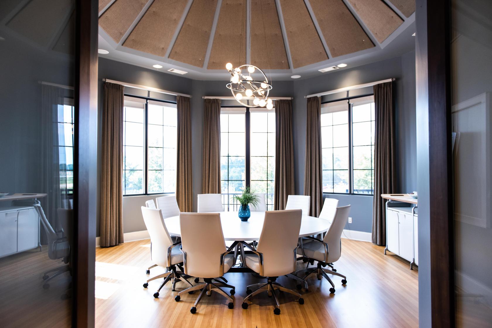 A meeting room with a round table, white chairs, large windows, and a modern chandelier at Paradigm Workhub.