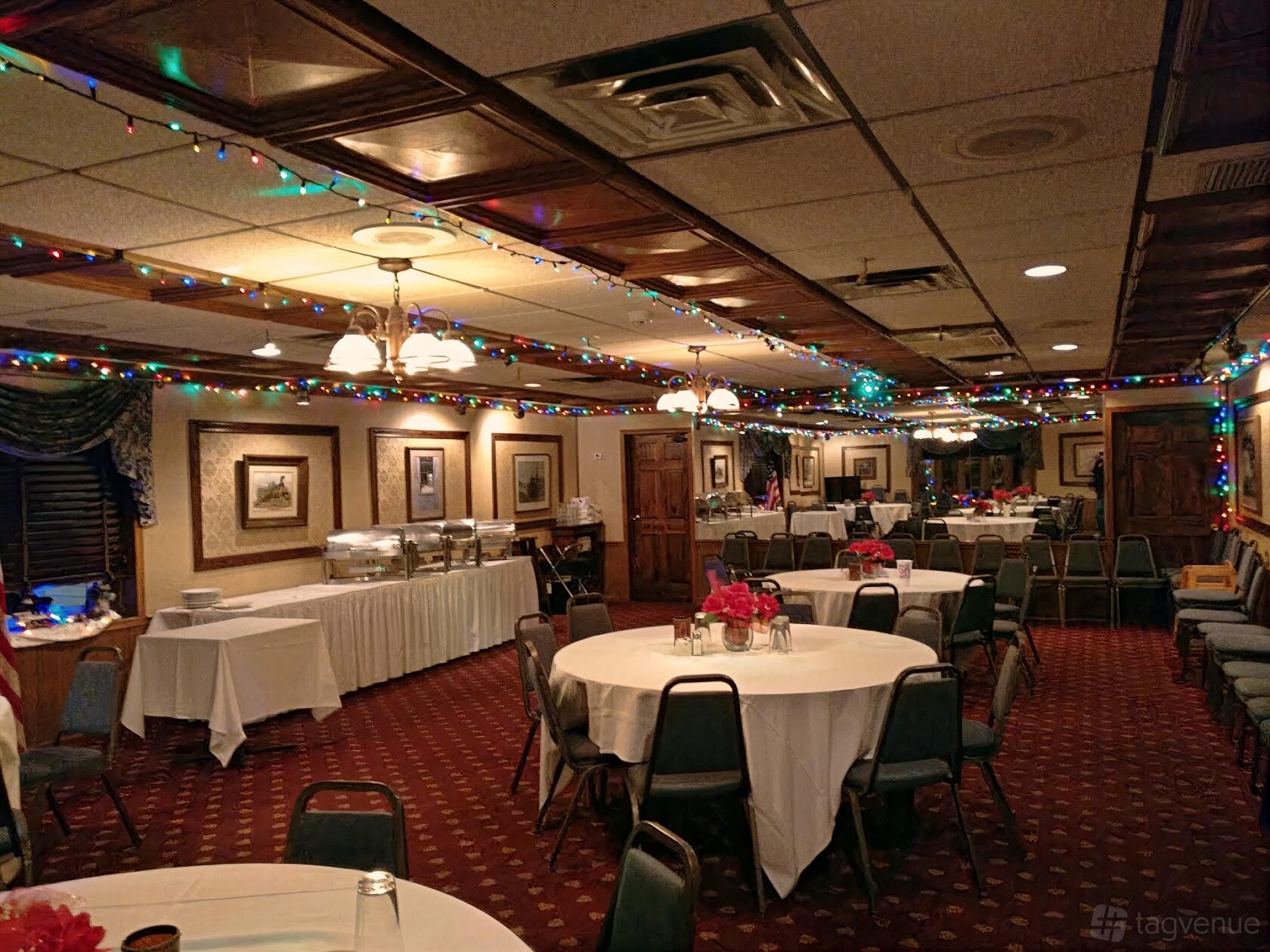 A dining room with round tables draped in white linens, red floral centerpieces, and string lights at Great Escape Restaurant.