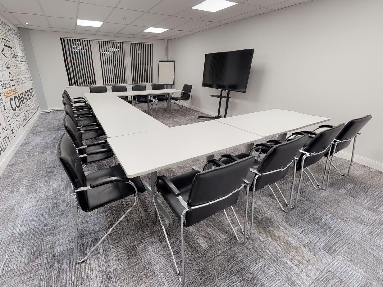 A meeting room with U-shaped white tables, black chairs, a large screen, and wall art at Portal Business Centre Dallam Court.