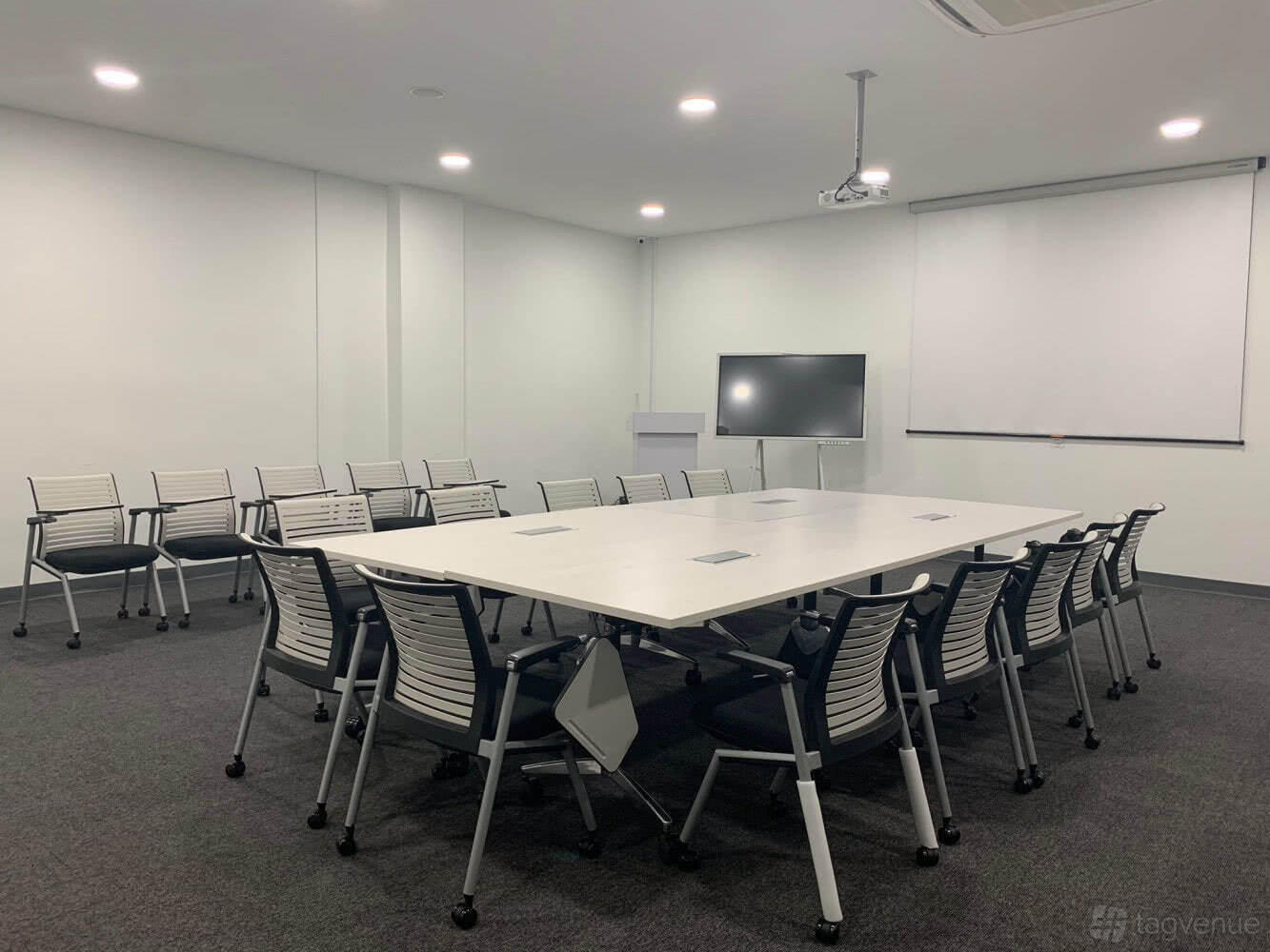 A training room with modular tables, ergonomic chairs, a ceiling projector, and a flat screen at The Training Room by CalGenie.