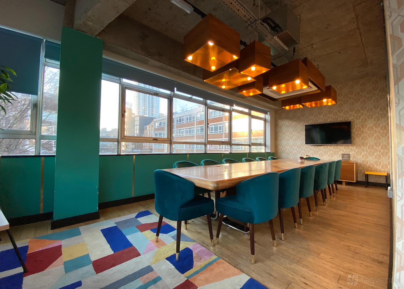 A meeting room with a long wooden table, teal chairs, geometric rug, and large windows at The Trampery Old Street.
