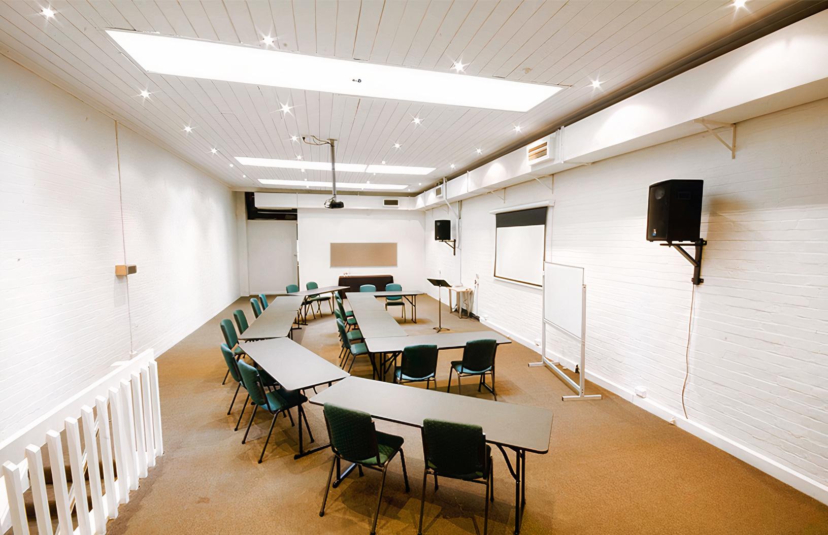 A meeting room with U-shaped tables, green chairs, a projector, and white brick walls at Melbourne City Conference Centre.