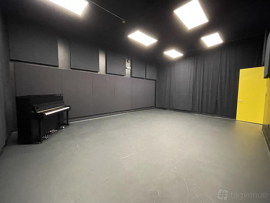 A rehearsal room with black acoustic wall panels, a black upright piano, and overhead fluorescent lighting at GARZA STUDIOS.