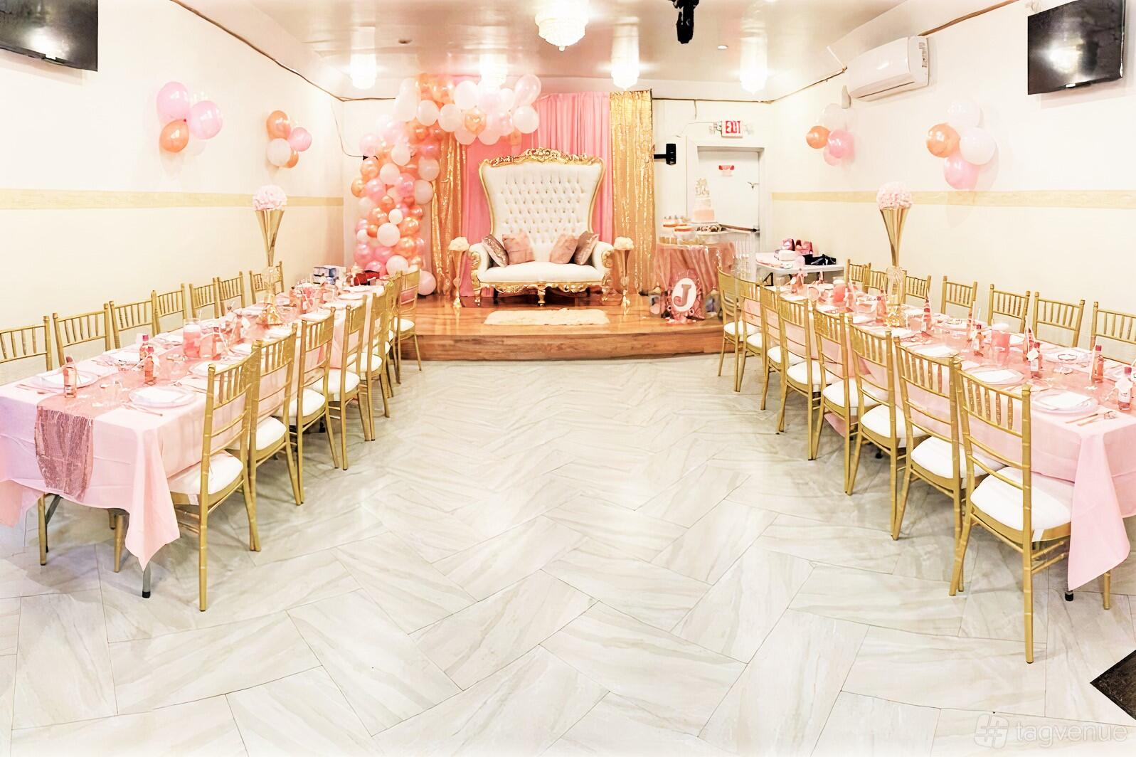 A hall with long banquet tables covered in pink linens, gold chairs, and a stage with a white tufted chair at La Belvedere LLC.