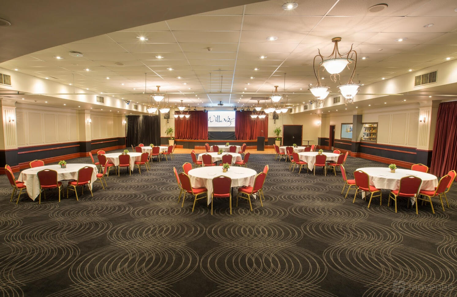 An event space with round banquet tables, red chairs, stage, and chandeliers at Village Green Hotel.