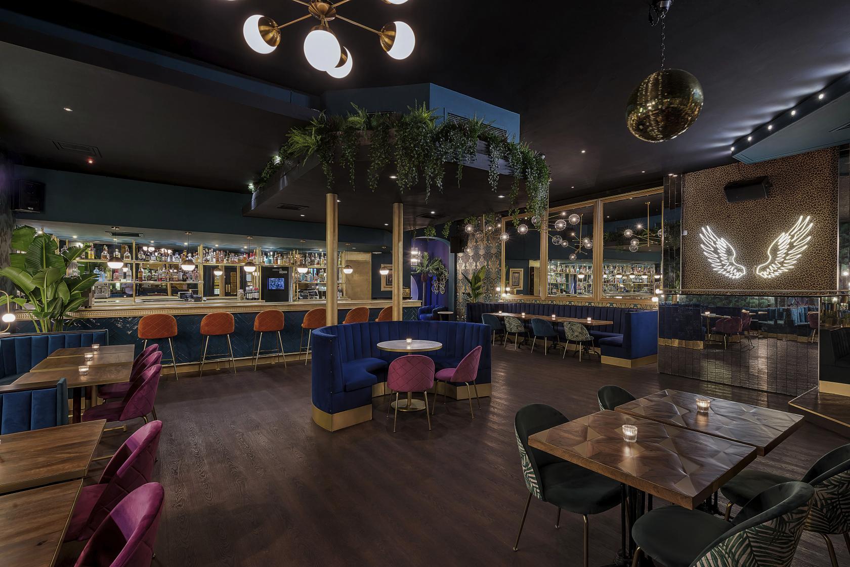 A cocktail bar with velvet seating, hanging greenery, and a mirrored wall with illuminated wings at Lost Society Battersea SW11.