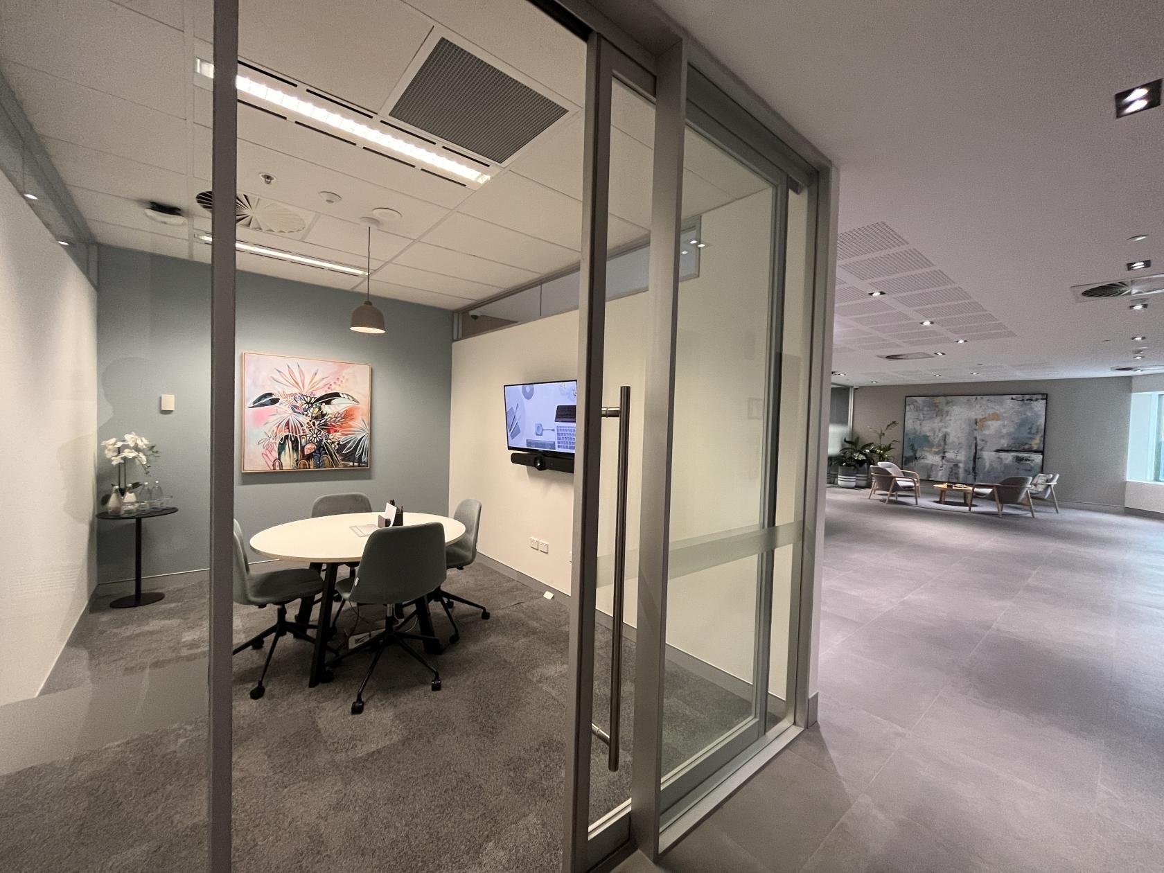 A meeting room with a round table, gray chairs, potted plants, and a wall-mounted screen at 108 St Georges Terrace.