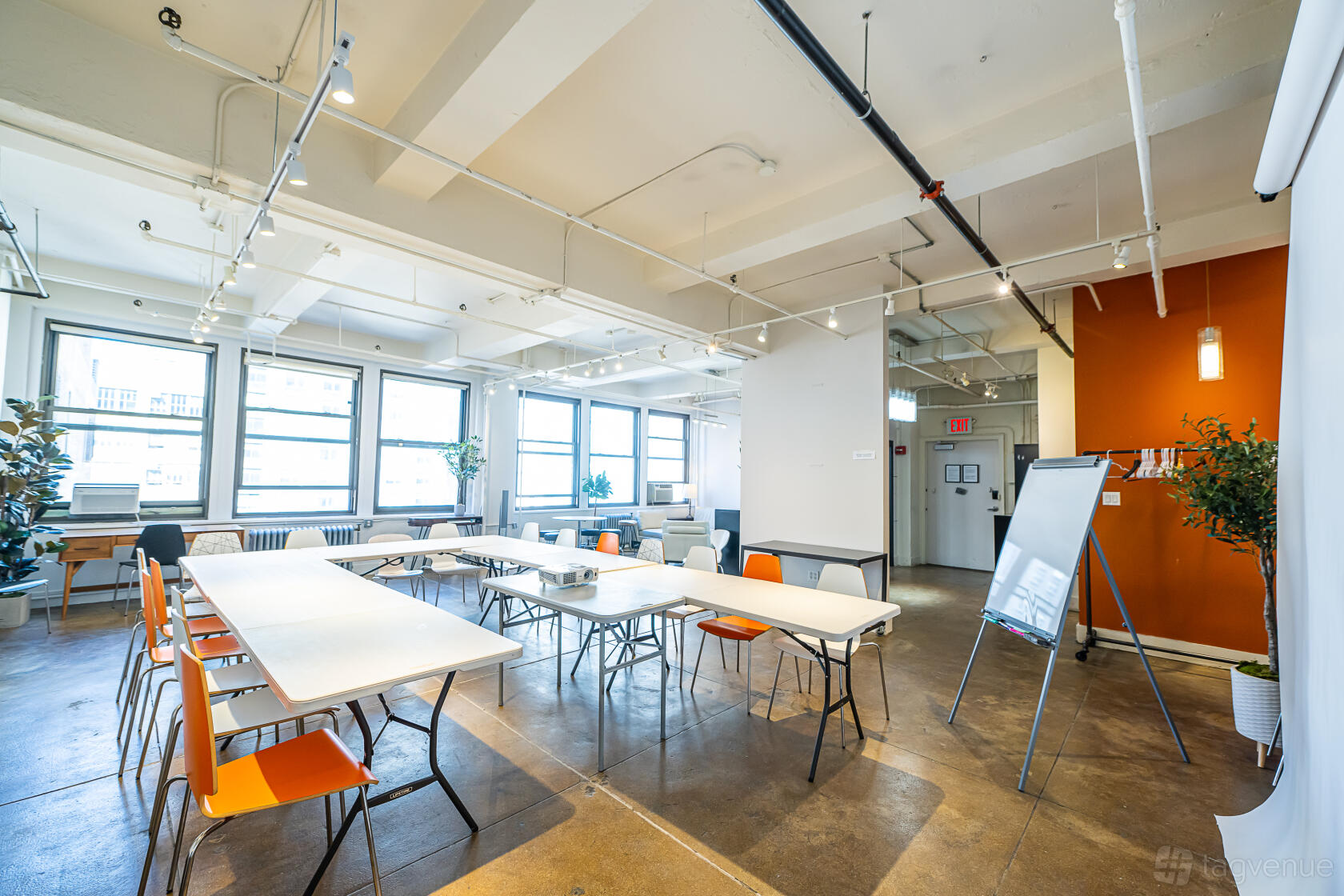 An event space with large windows, white tables arranged in a U-shape, and orange chairs at Studio 1418.