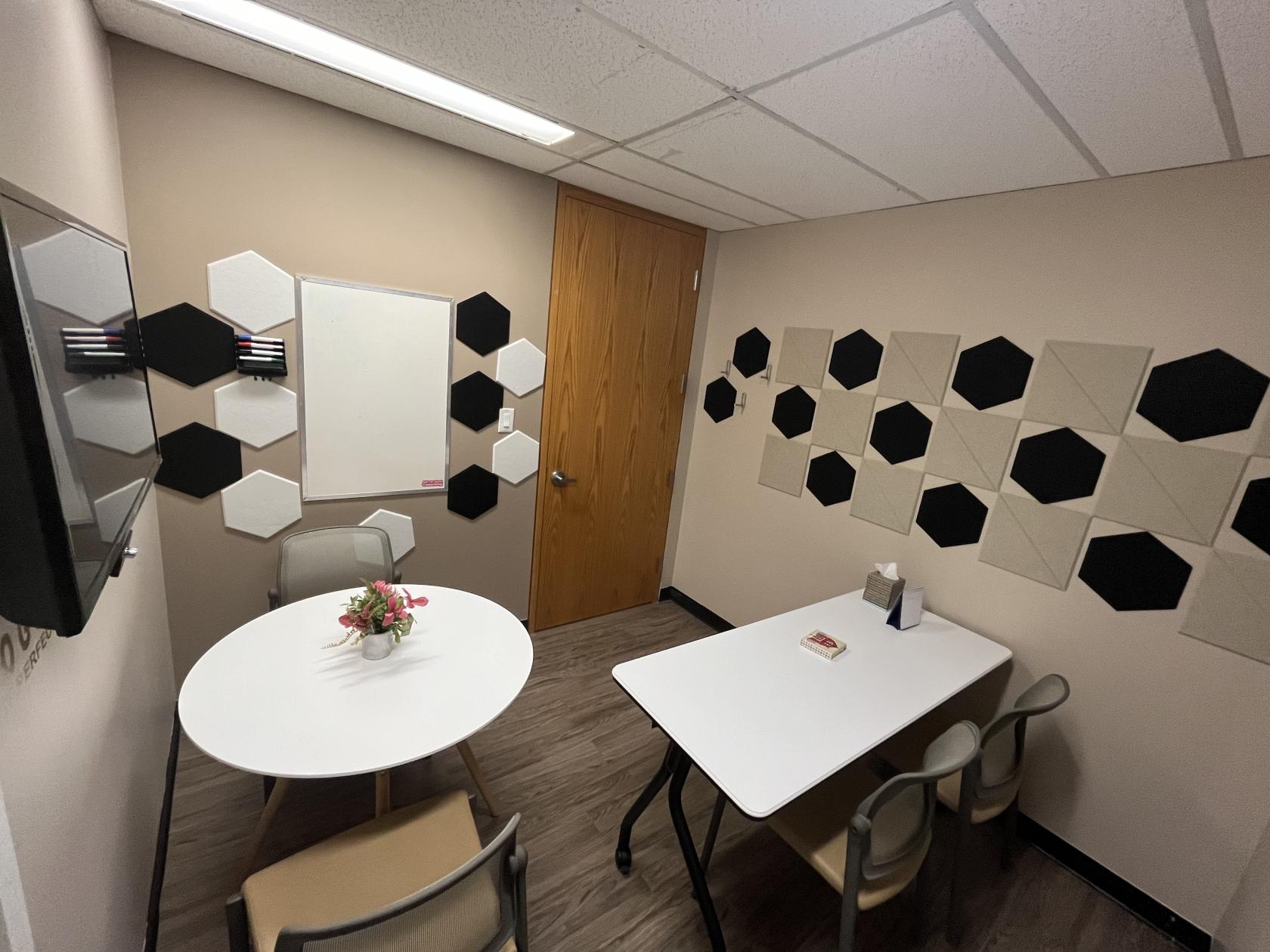 A meeting room with acoustic hexagon wall panels, a round table with a flower arrangement, and a whiteboard at AHANSA Business Center.