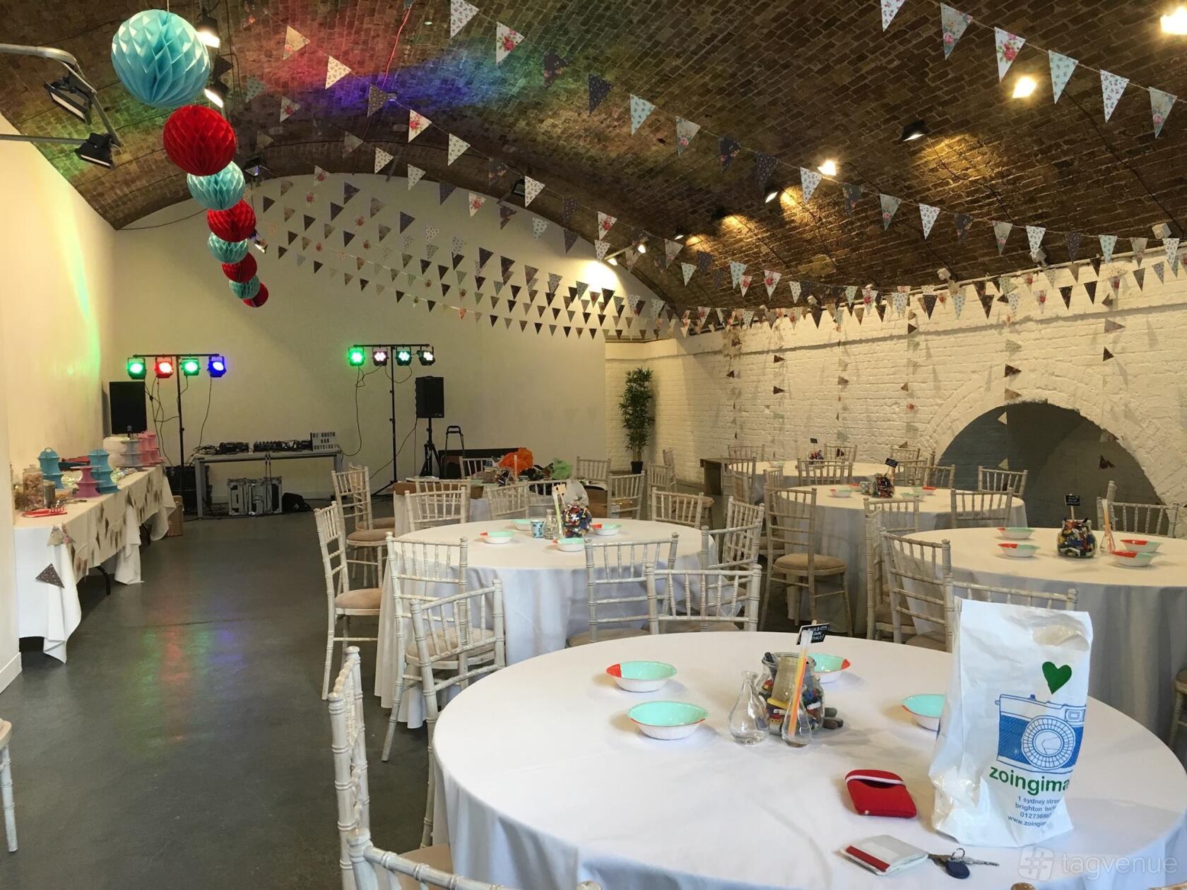 An event venue with exposed brick vaulted ceilings, round tables, white chairs, and bunting decorations at Hoxton Arches.