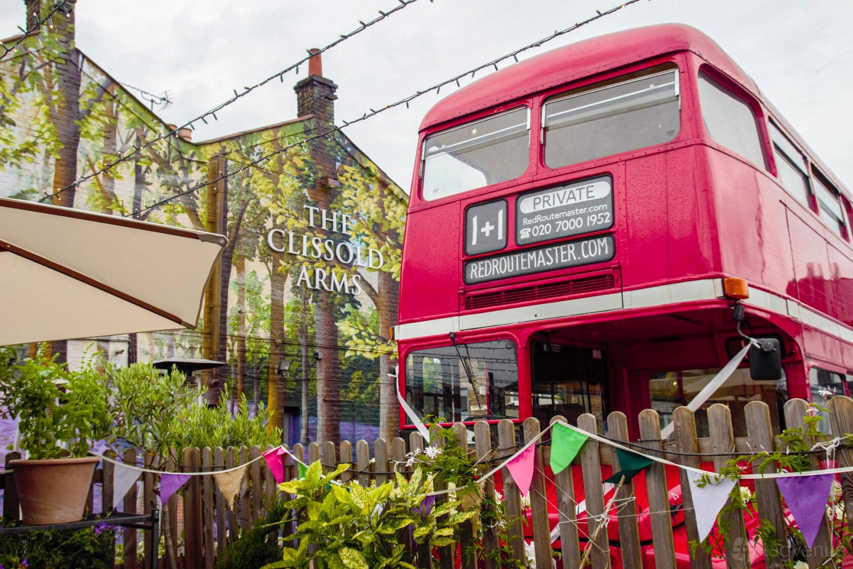 A pub with a mural-covered exterior, festoon lighting, bunting, and a vintage red double-decker bus at Clissold Arms.