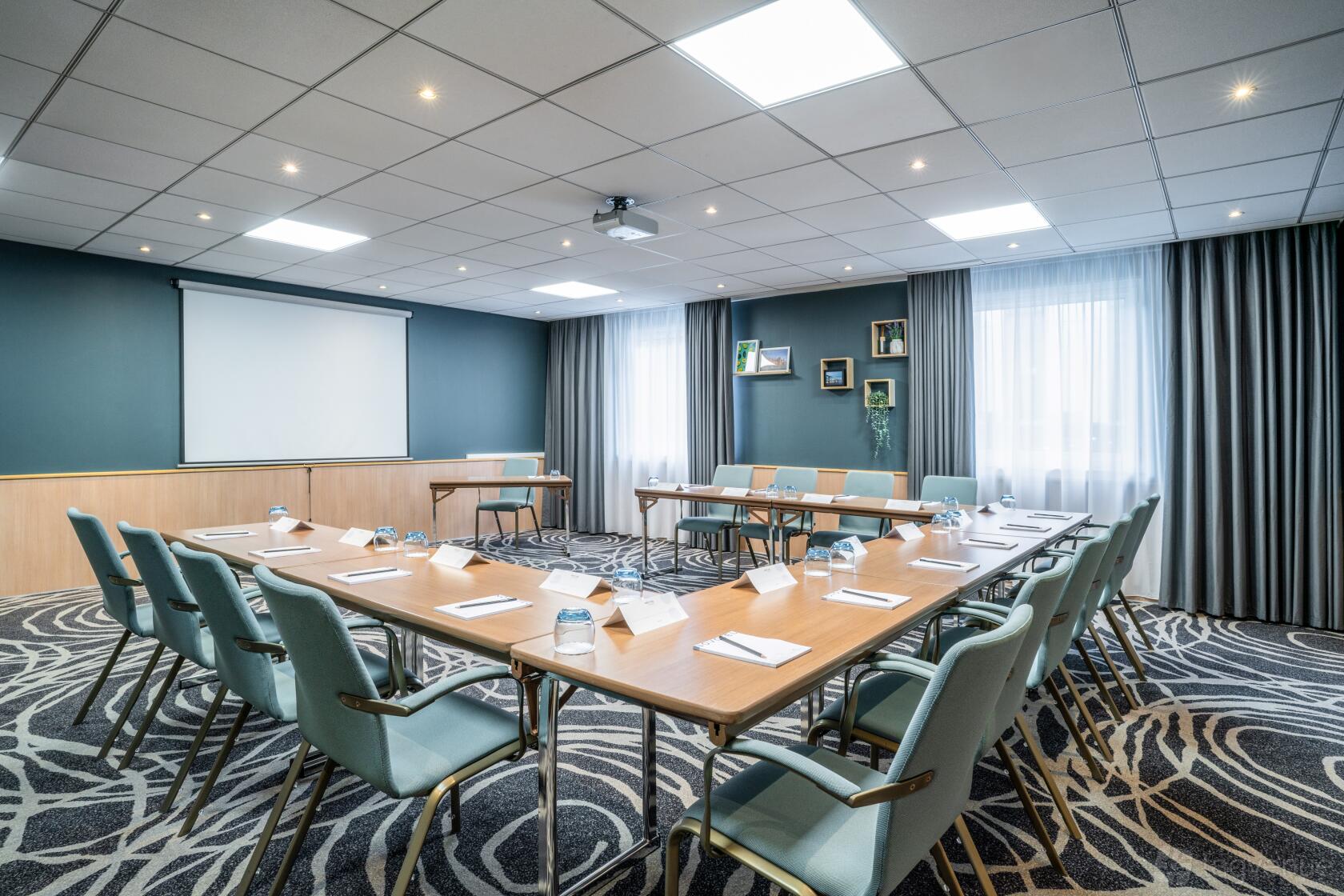 A meeting room with U-shaped tables, padded chairs, a ceiling projector, and large curtained windows at Novotel Glasgow Centre.