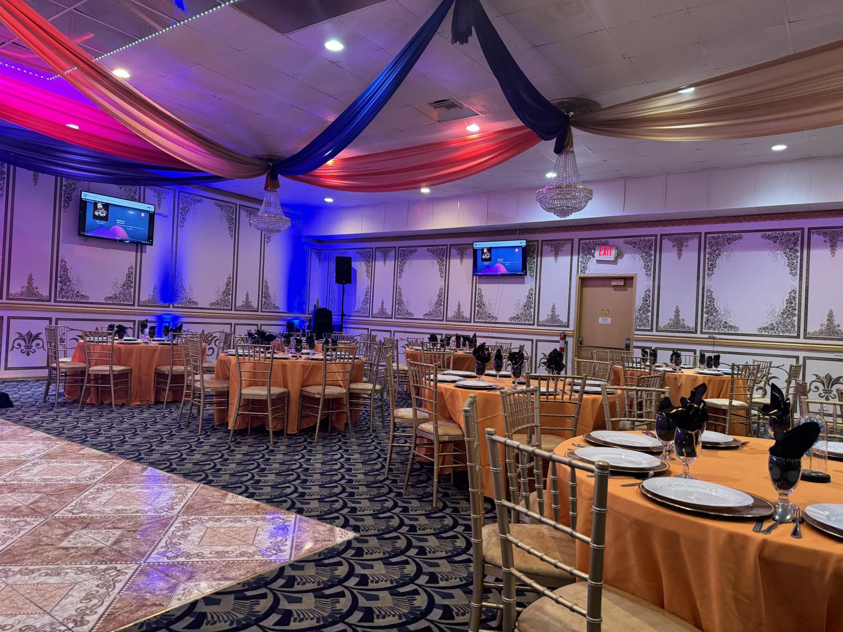 A banquet hall with chandeliers, draped ceiling fabric, round tables with orange linens at Carmelle Banquet Hall.