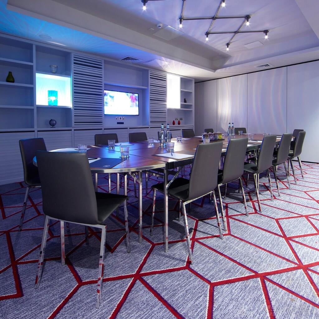 A function room with a U-shaped meeting table, black chairs, built-in shelving, and a large screen at Malmaison Glasgow.