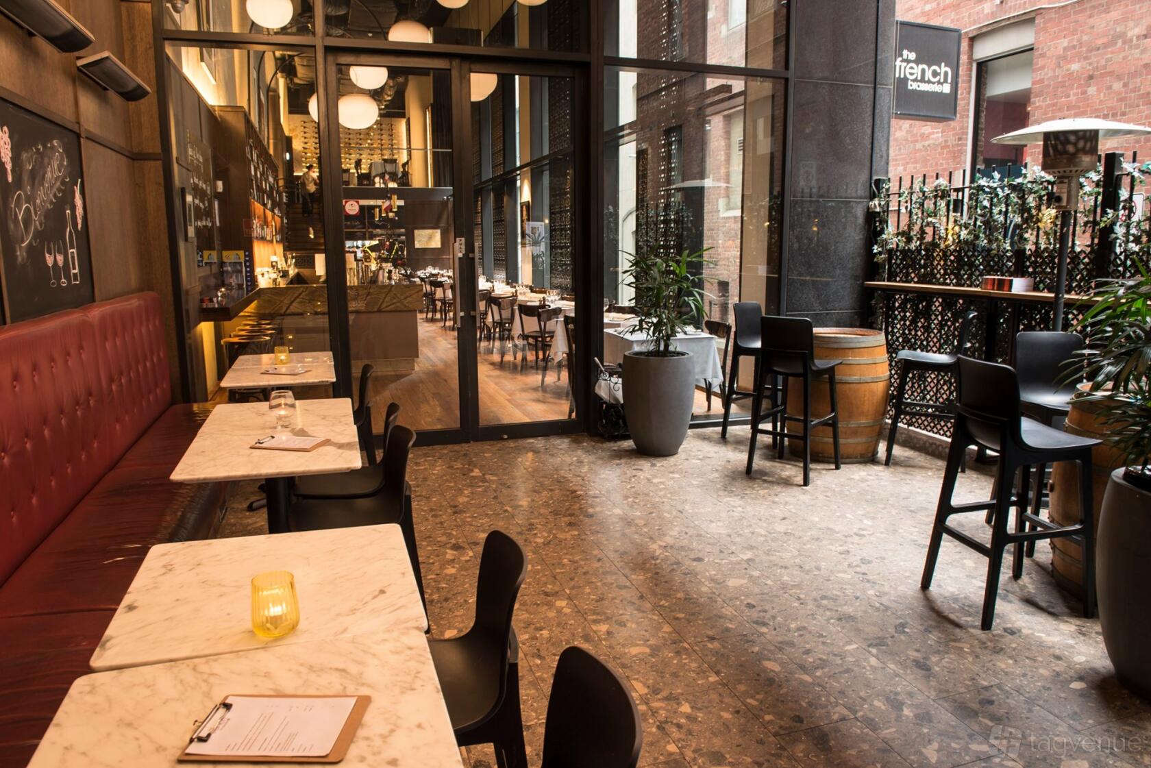A restaurant terrace with marble tables, cushioned banquette seating, and potted plants at The French Brasserie.