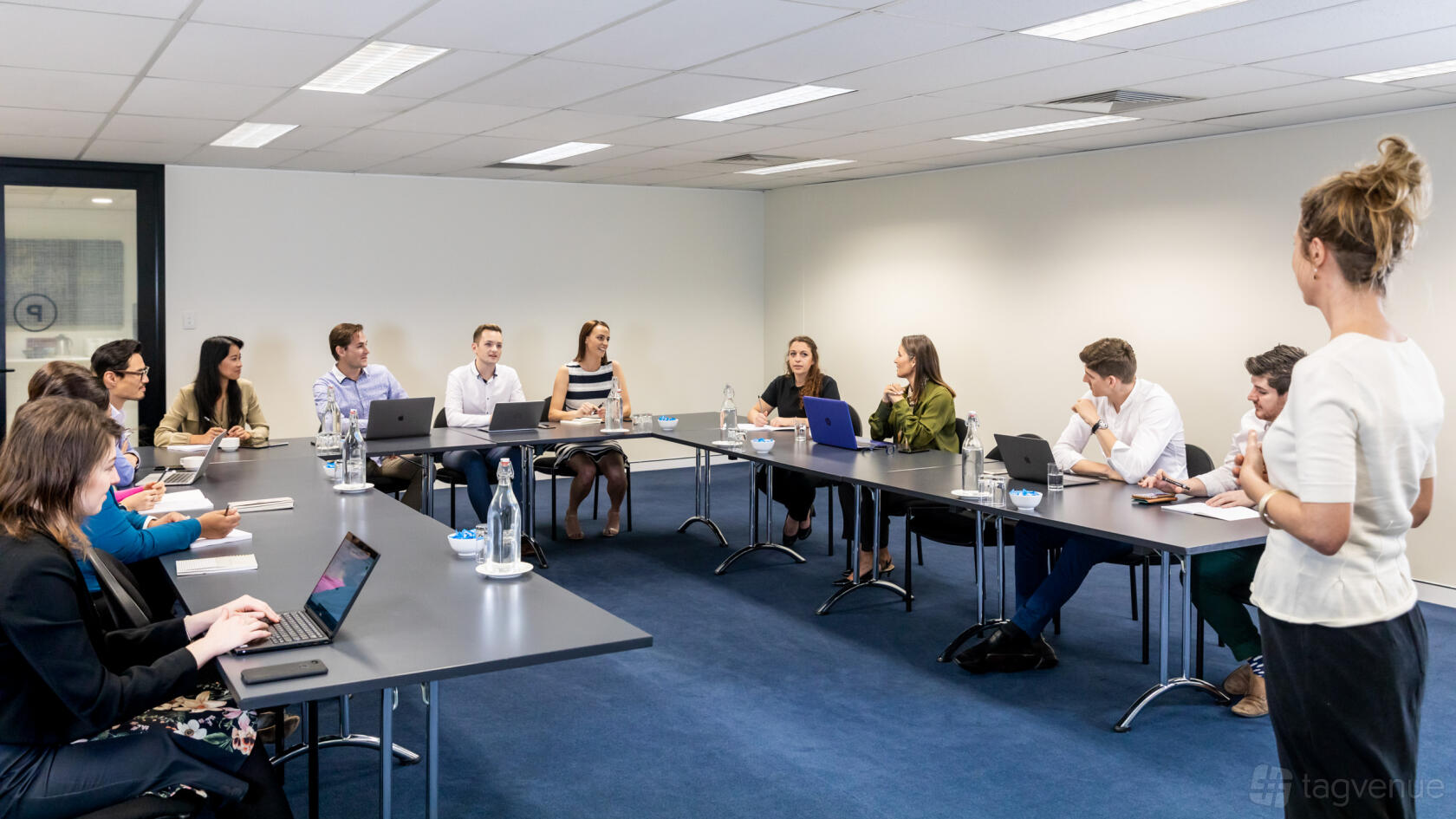 A coworking conference room with U-shaped tables, blue carpet, and ceiling panel lighting at Christie Spaces Conferencing Brisbane.