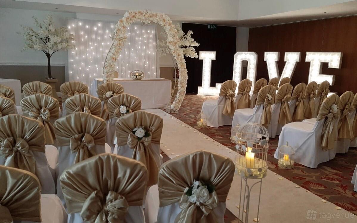 An event space with gold chair sashes, floral arch, marquee LOVE letters, and candle lanterns at Hellaby Hall Hotel.