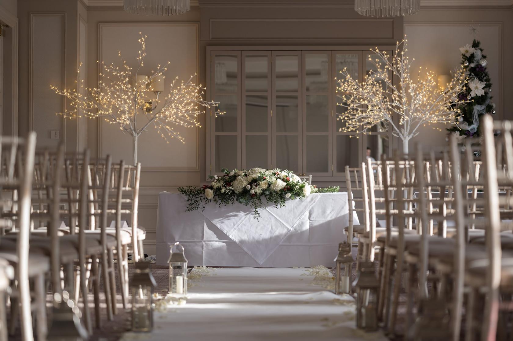 A hotel ceremony space with rows of wooden chairs, floral centerpieces, and decorative lighted trees at Down Hall Hotel.